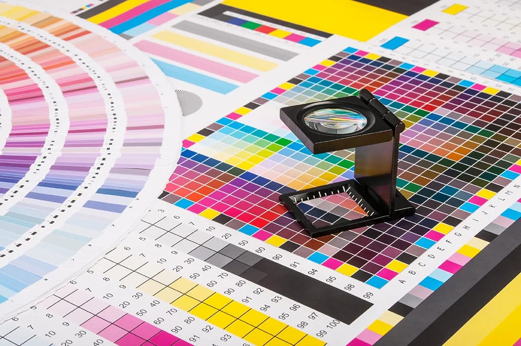 A magnifying glass sits on a colourful print test sheet filled with various colour swatches, gradients, and grids, commonly used in graphic design and printing quality control.
